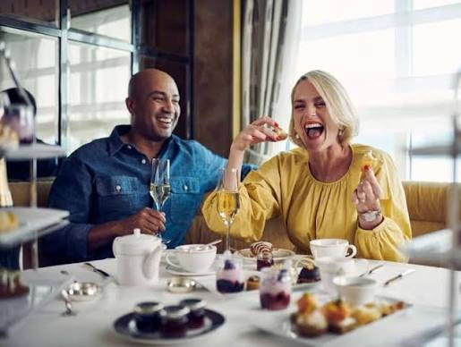 A couple enjoys a meal, laughing over champagne in a bright, elegant restaurant. The woman wears yellow, the man in denim. Various dishes on the table.