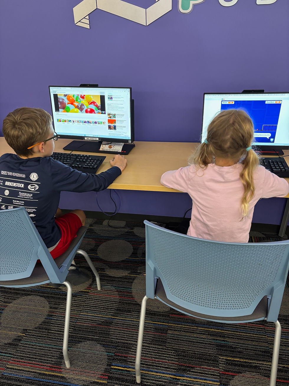 Two kids using computers in a room with a purple wall and colorful carpet. One screen shows balloons, the other a blue grid.