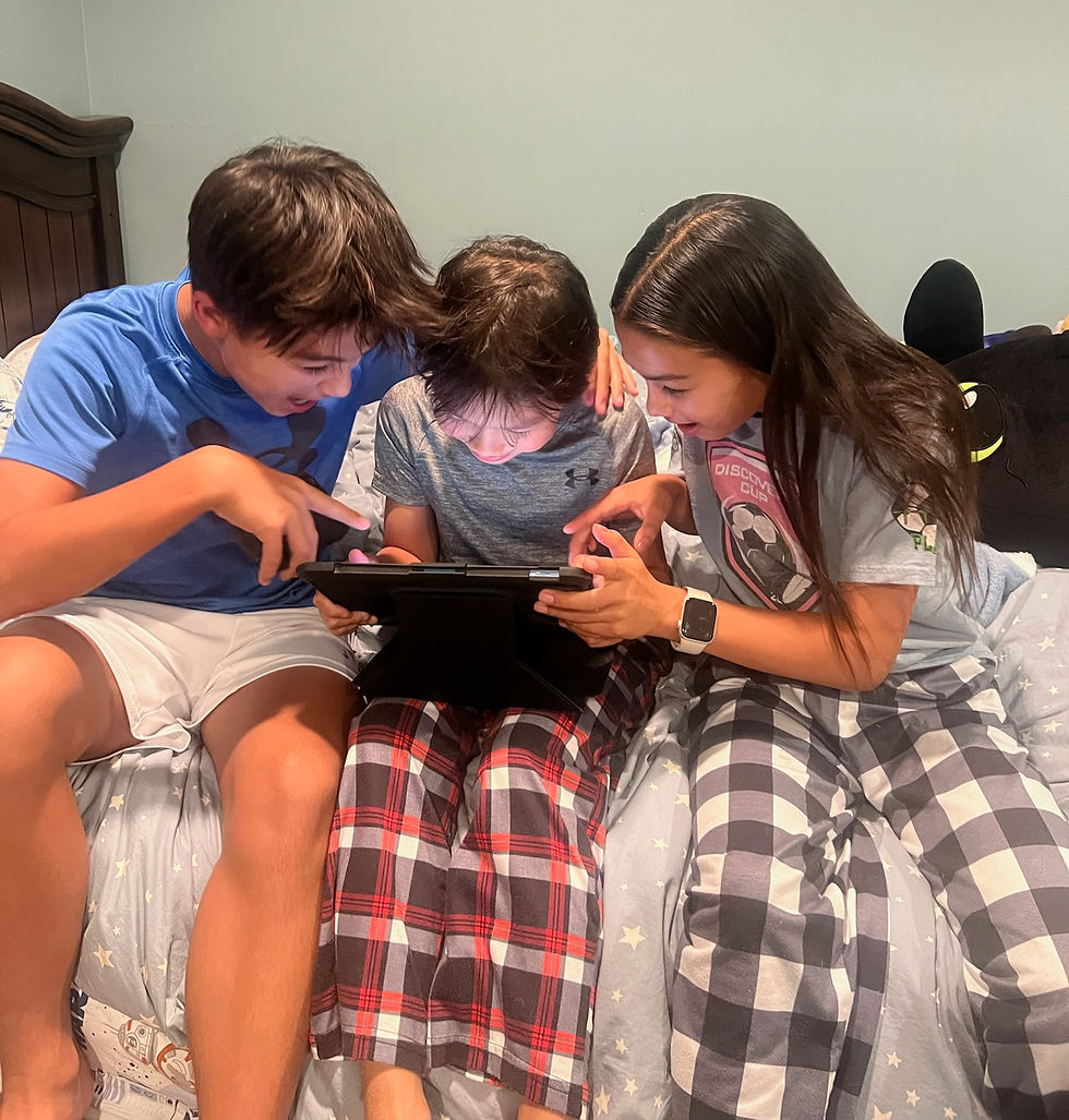 Three children in pajamas sit on a bed, intently looking at a tablet. The room has blue walls and a starry comforter.