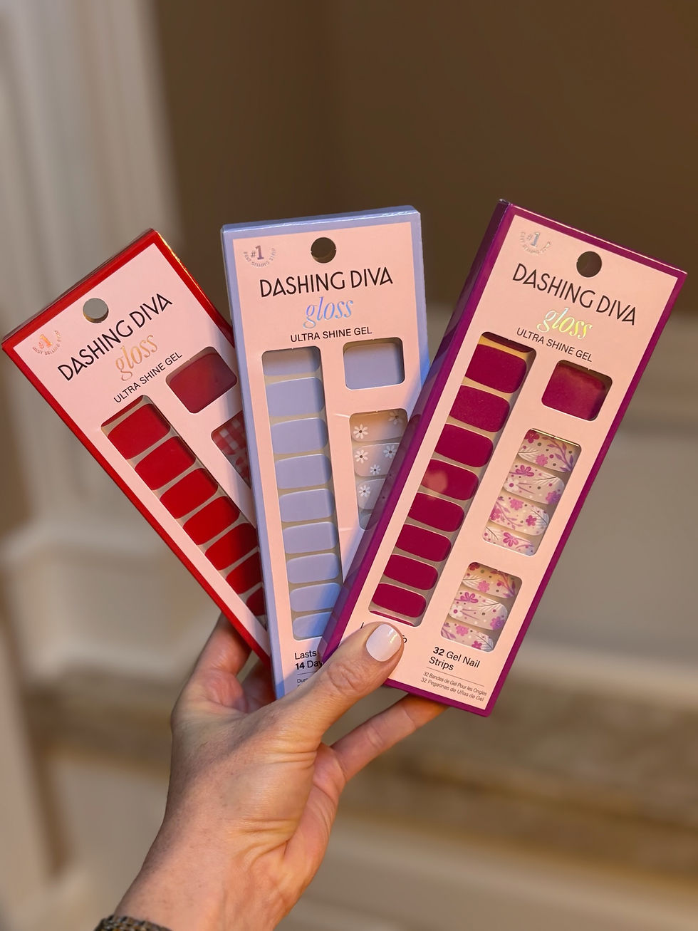A hand holds three packs of Dashing Diva nail strips in red, blue, and floral patterns. Text reads "gloss" and "Ultra Shine Gel" on packaging.