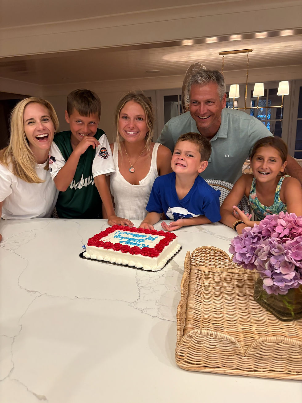 Family of six smiling around a cake with red icing. Bright kitchen setting, purple flowers, wicker tray on marble counter. Warm mood.