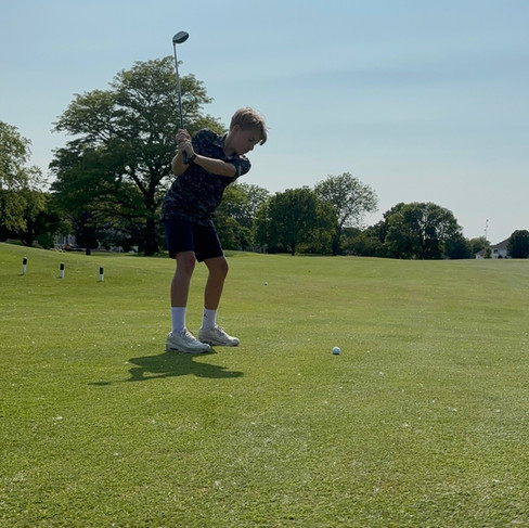 Young golfer swinging club on lush green course under clear sky, trees in background, wearing dark clothes and white shoes.