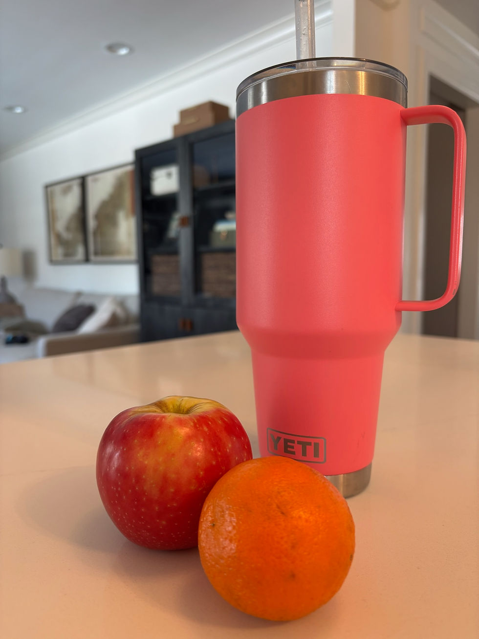 A pink Yeti tumbler, red apple, and orange on a kitchen counter. Background shows framed maps and shelves. Bright and cozy setting.