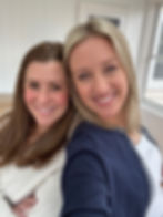 Two smiling women pose for a selfie indoors by a bright window, one in a navy cardigan and one in a cream sweater.