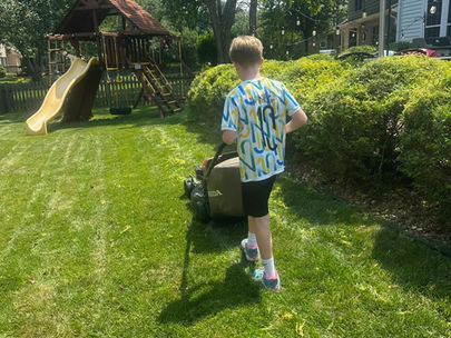 Boy mowing a sunny backyard lawn with a Honda mower near a wooden playset, trees, and a house with string lights.