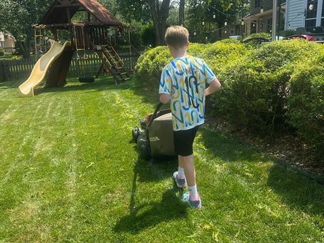 Boy mowing a sunny backyard lawn with a Honda mower near a wooden playset, trees, and a house with string lights.