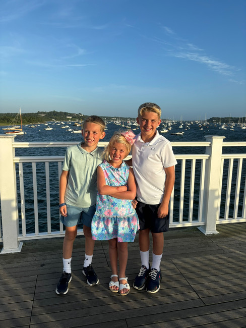 Three smiling children pose on a boardwalk by the sea. Boats are visible in the background. The sky is clear and blue.