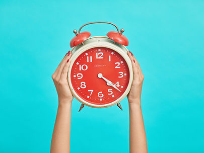 Hands with red nails holding a vintage red alarm clock against a bright turquoise background. The clock reads 10:09.