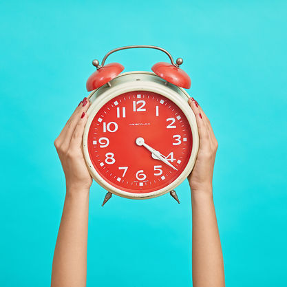 Hands with red nails holding a vintage red alarm clock against a bright turquoise background. The clock reads 10:09.