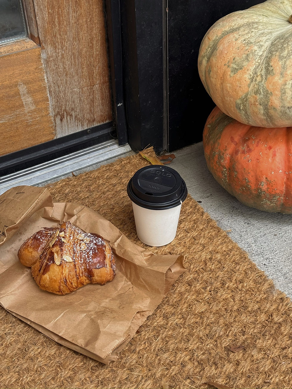 Croissant and coffee sit on a brown paper bag atop a woven mat. Two pumpkins rest nearby against a wooden door, creating an autumn vibe.