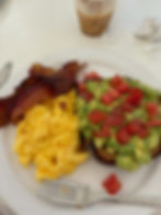 Plate with bacon, scrambled eggs, avocado toast topped with tomatoes. Iced coffee in background. Fork and crumpled napkin on white table.