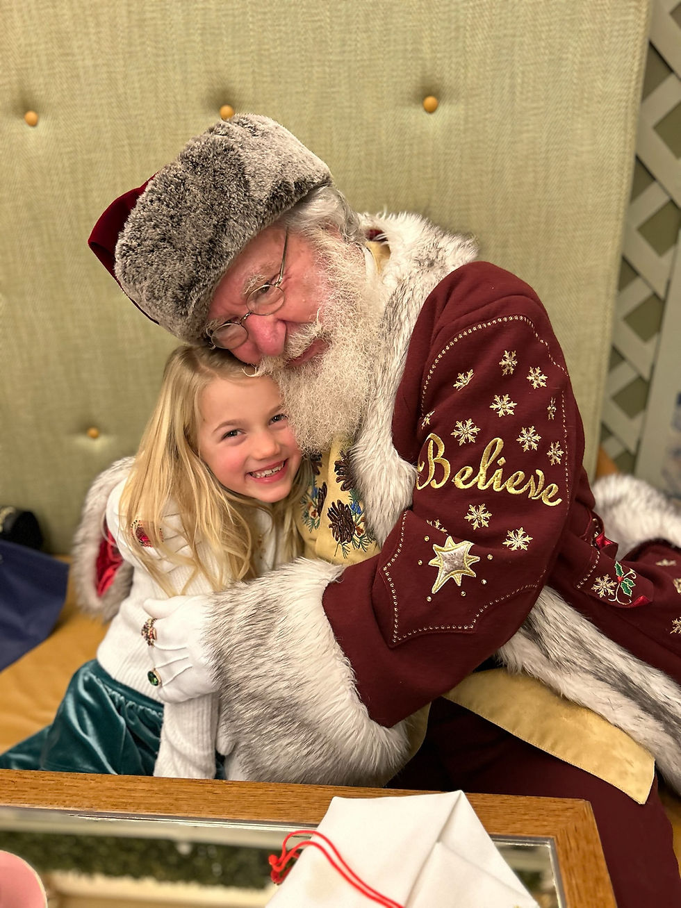 Santa in a festive red coat hugs a smiling child on a tufted bench. The word "Believe" is on his sleeve. Warm and joyful mood.