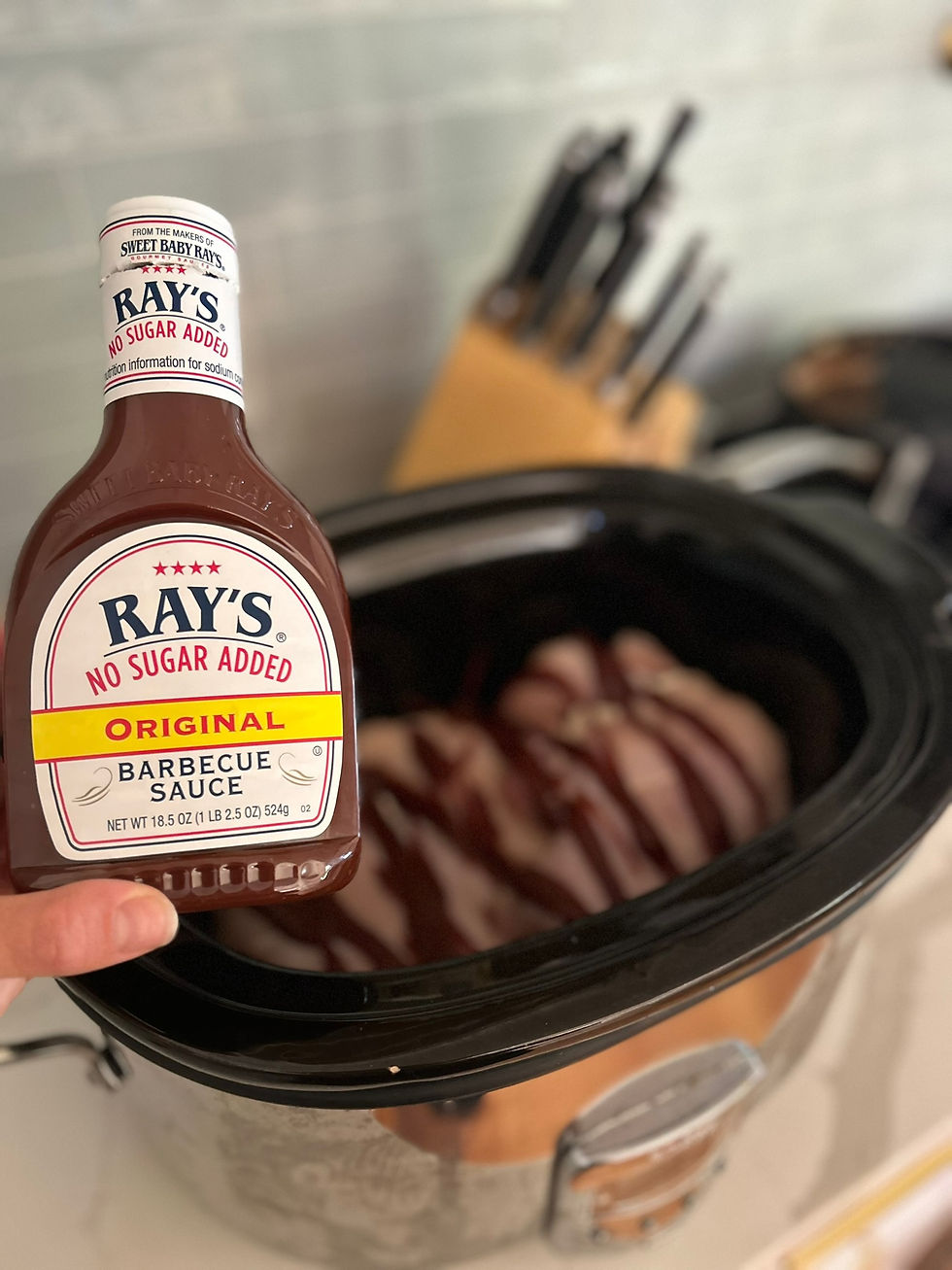 Hand holding Ray’s No Sugar Added Original Barbecue Sauce in front of a slow cooker filled with ribs and a knife block in back.