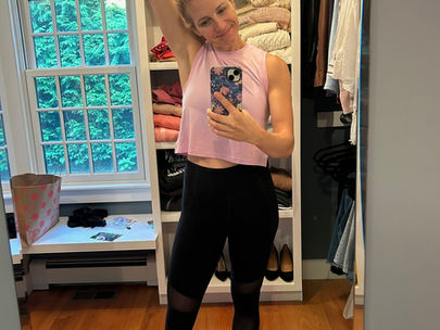 Woman in pink top and black leggings takes a mirror selfie in a walk-in closet. She smiles with one arm raised. Shelves hold clothes and hats.