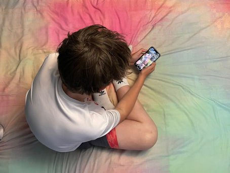 Child in white shirt sits cross-legged on a rainbow bedspread, engrossed in a smartphone showing colorful content. Cozy atmosphere.