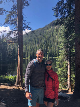 Two people smiling in a forest setting near a lake. One wears a red jacket, the other in a striped shirt. Tall trees and clear blue sky.