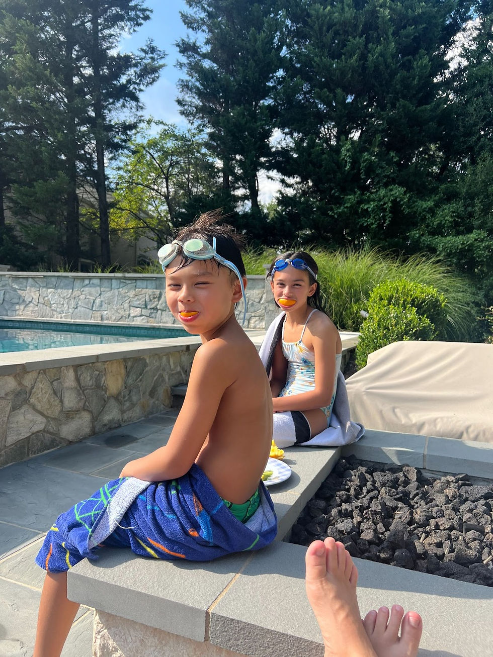 Woman wearing sunglasses smiles by colorful mosaic wall, with terracotta buildings and greenery in the background. Bright, relaxed setting in Barcelona. 