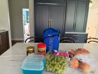 A blue lunch bag, peanut butter, jelly, grapes, apples, bread, and a food container on a marble kitchen counter. Dark cabinets in the background.