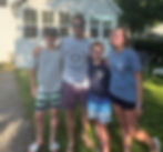 Smiling man and three kids stand outside a white house, wearing Delavan Lake Marina shirts, in warm afternoon light.