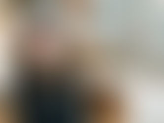 Smiling woman with red glasses sits on a brown leather chair, with a colorful abstract patterned wall in the background.