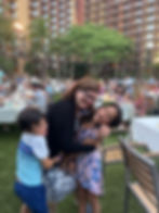 A woman hugs two laughing children on a grassy area with tables and a large building in the background. Bright and joyful atmosphere.