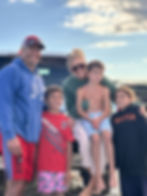 Smiling family of five posing by a pickup truck on a beach; kids in sports hoodies and red shirt, cloudy sky overhead.