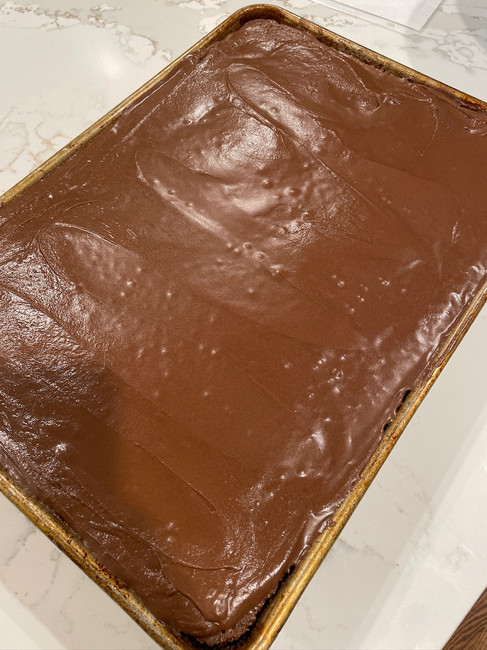 A sheet pan with smooth chocolate frosting on a cake, set on a marble countertop. The surface is glossy and rich in color.