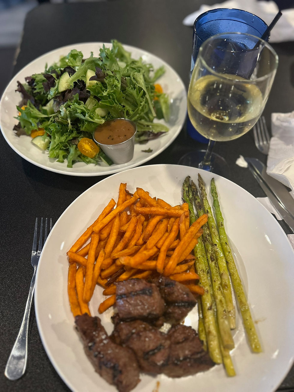 Plates of steak bites, sweet potato fries, asparagus, and salad sit on a dark table. A glass of white wine and a blue tumbler are visible.