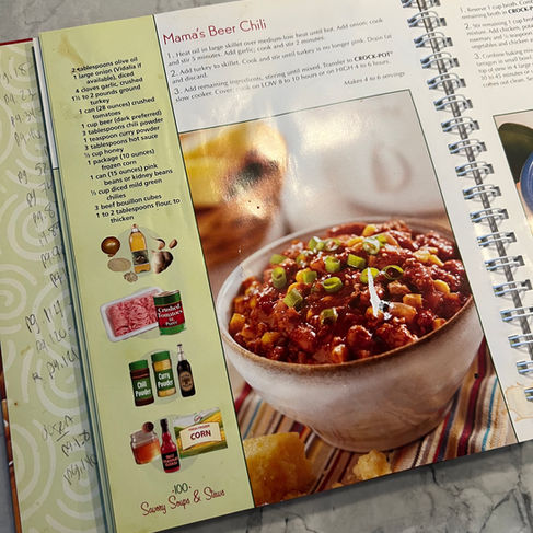 Open cookbook on a marble counter, featuring "Mama's Beer Chili" recipe with ingredients, instructions, and a bowl of chili on a striped surface.