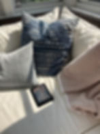 Cozy sunlit patio daybed with striped and cream pillows, a blanket, and a Kindle showing It’s a Love Story.