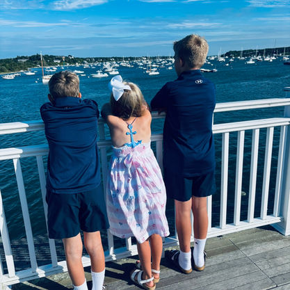 Three children in navy clothes and a pink dress stand on a dock, leaning on a white railing, looking over boats on a sunny day.