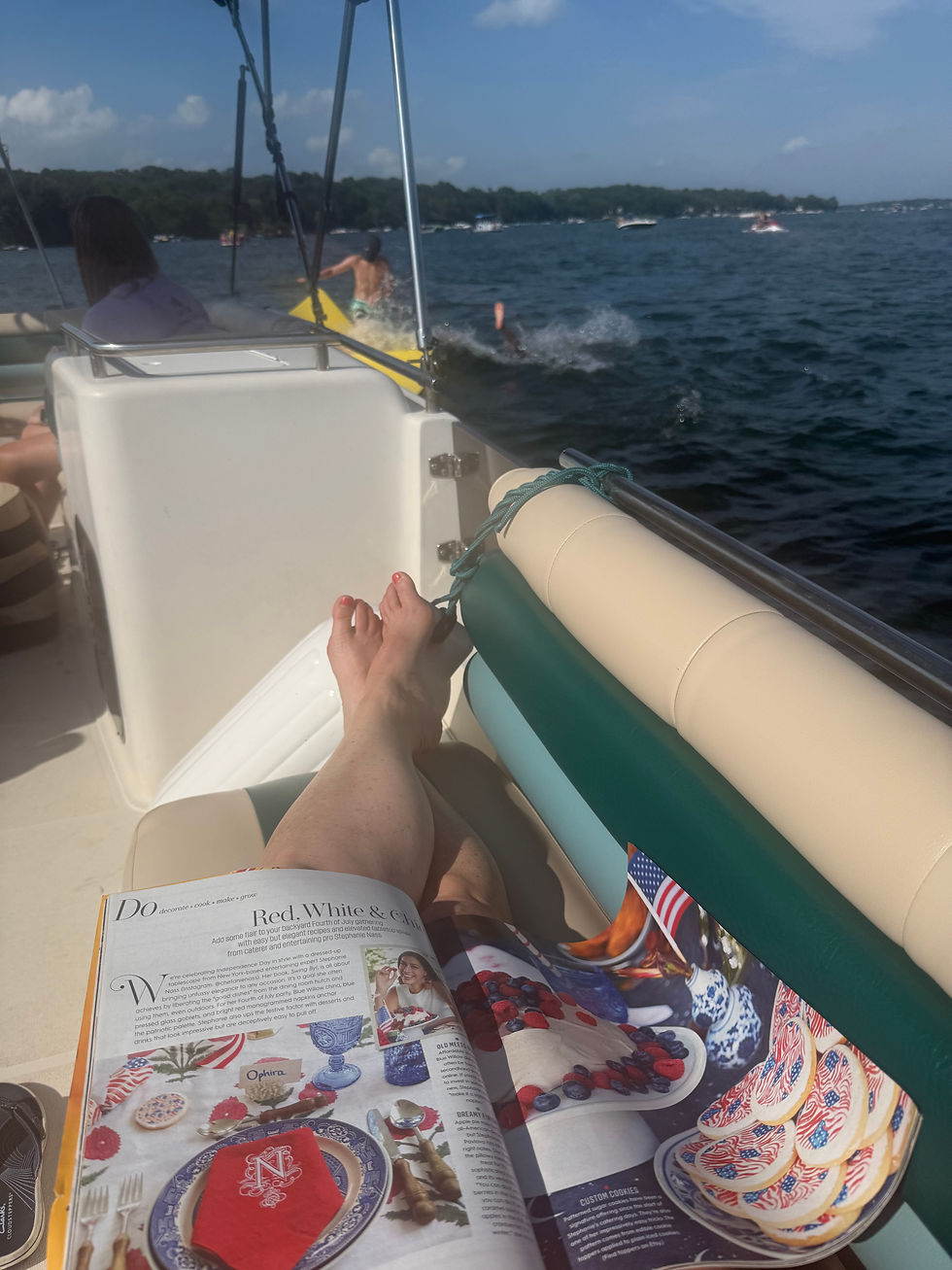 Feet on a boat beside an open magazine, while a swimmer splashes nearby on a sunny lake.