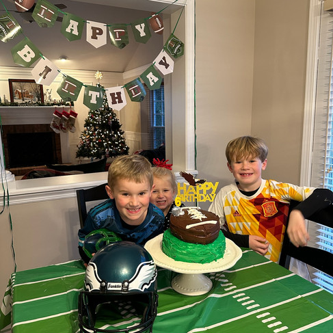 Three smiling kids around a football-themed cake with "Happy 6th Birthday" topper, set on a matching tablecloth. Christmas tree and decor in the background.