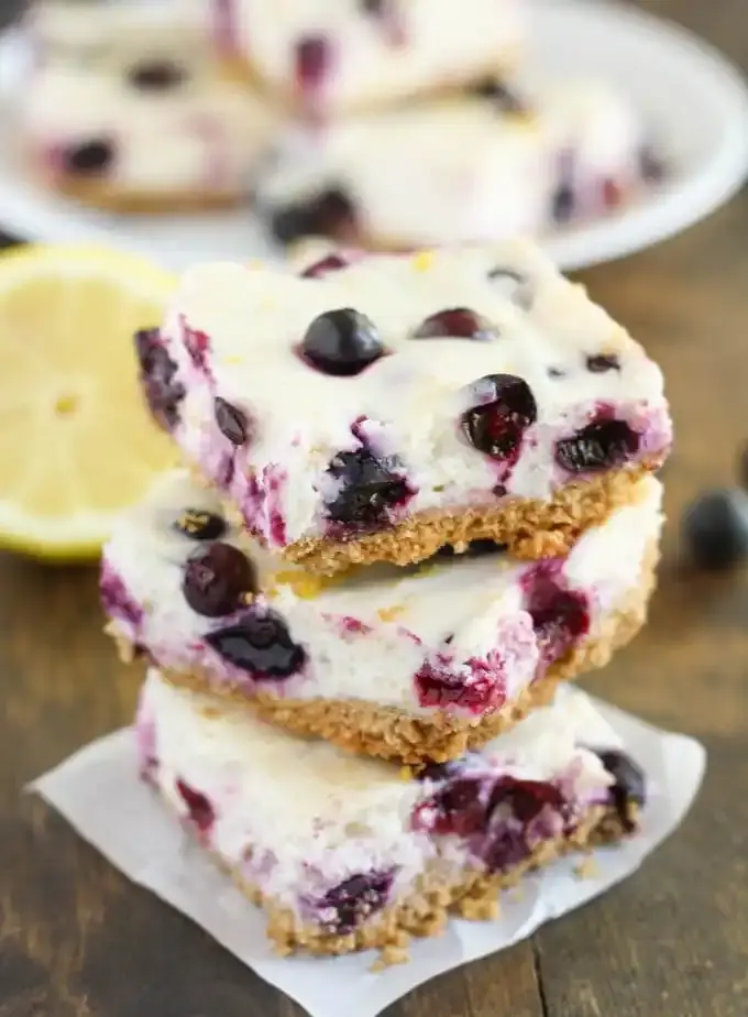 Stack of three blueberry cheesecake bars with a crumbly crust on a wooden table. A lemon slice and a blurred plate are in the background.