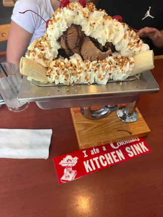 Banana split with chocolate ice cream, whipped cream, and cherries on a table. A sign reads, "I ate a Colossal KITCHEN SINK.”
