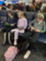 Three kids seated at an airport gate, using tablets. Backpacks on floor, passengers in the background. Casual setting with focused mood.