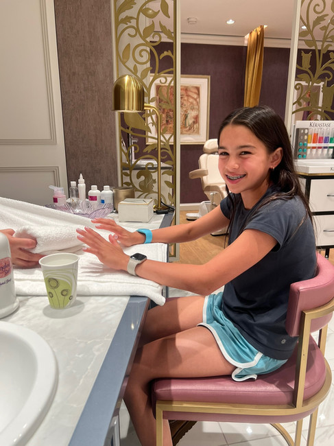 Girl smiling during a manicure at a salon. She's seated on a pink chair, wearing a dark shirt and blue shorts. Gold decor and beauty items surround her.