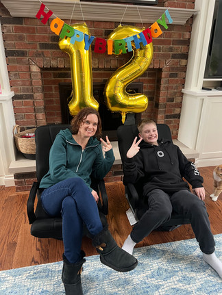 A woman and boy flash peace signs in chairs, smiling. Gold "12" balloons and "Happy Birthday" banner hang in brick fireplace background.