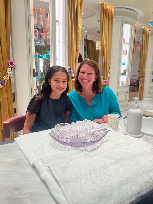 Smiling woman and girl seated at a table with a purple glass bowl and towels. Background shows shelves with products and golden curtains.