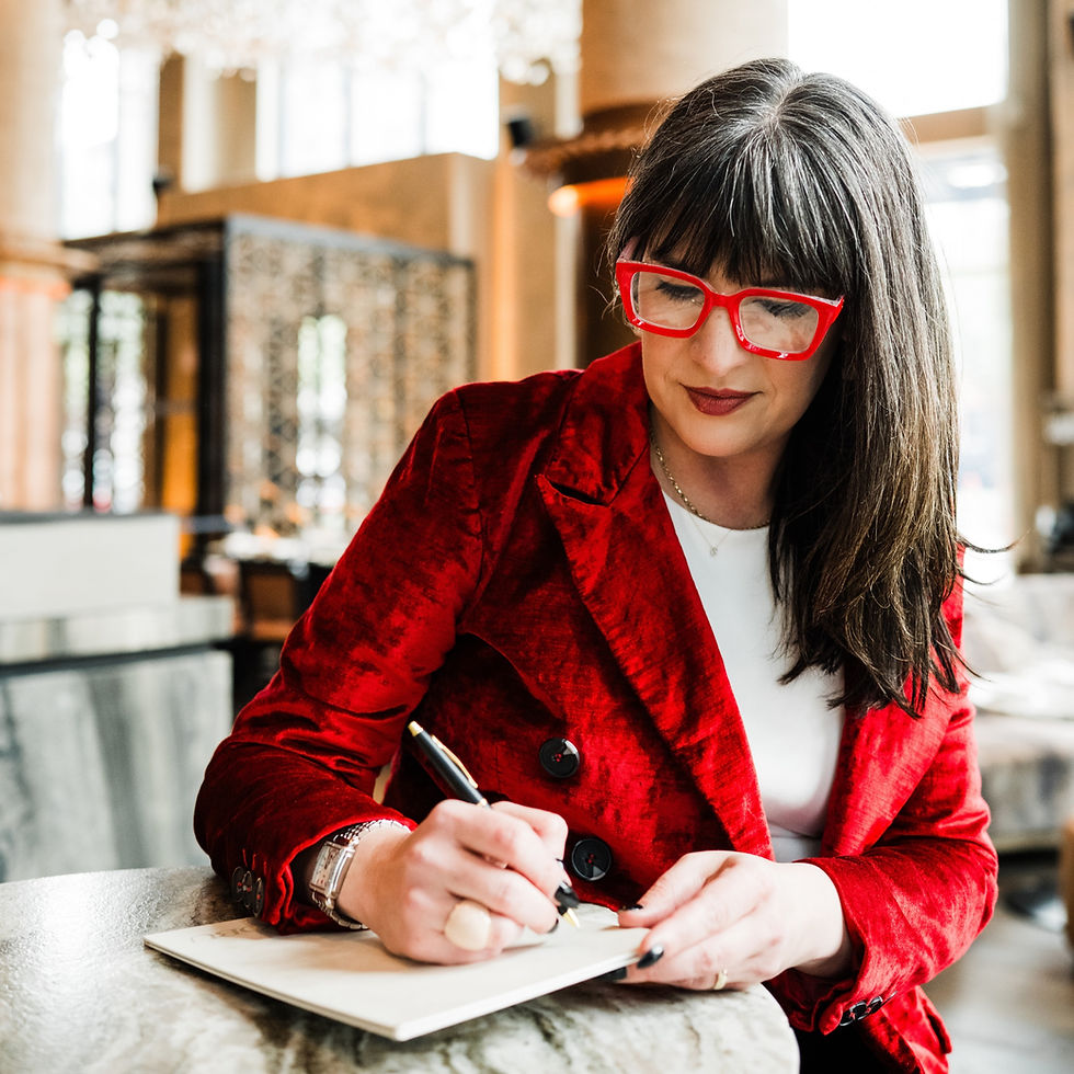 Woman in a red jacket and glasses writing in a notebook. She's seated in a stylish indoor setting with soft, natural light.