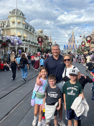 Family posing happily on a busy street with festive decorations. Disney castle in the background. Bright clothing, balloons, and a lively atmosphere at Magic Kingdom.