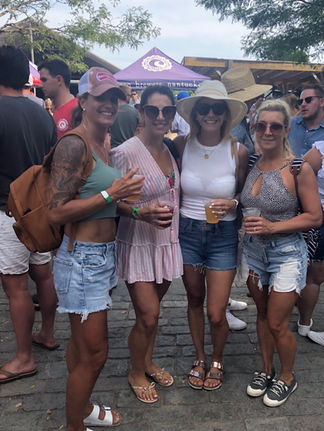 Four women smiling, holding drinks at an outdoor event. They wear summer outfits. A "brewery" tent and trees are in the background.