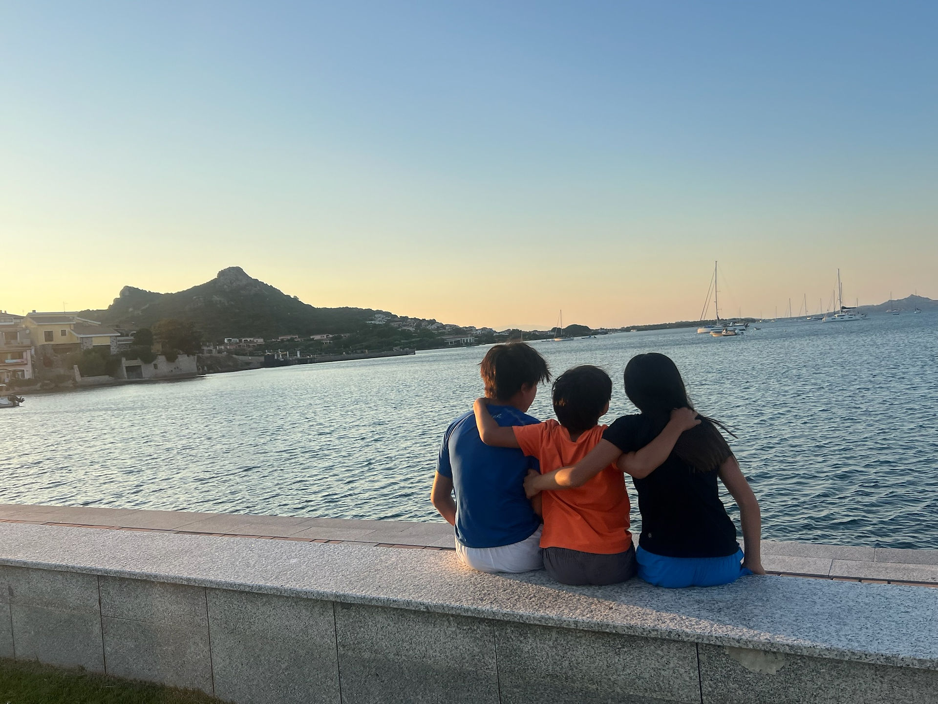 Three people sit on a waterfront ledge, arms around each other, watching a calm sea with sailboats at sunset. A mountain is in the background.