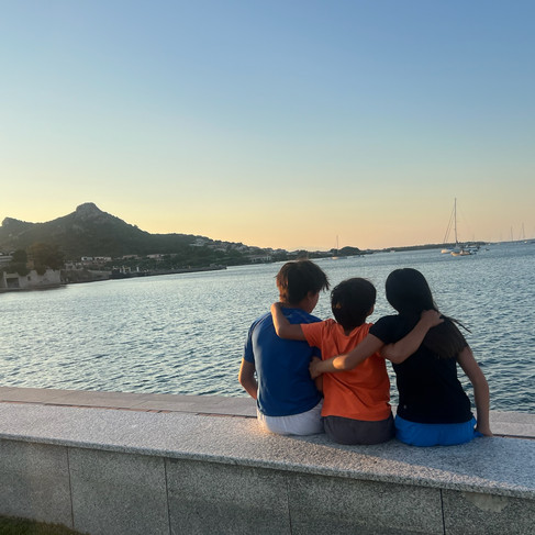 Three people sit on a waterfront ledge, arms around each other, watching a calm sea with sailboats at sunset. A mountain is in the background.