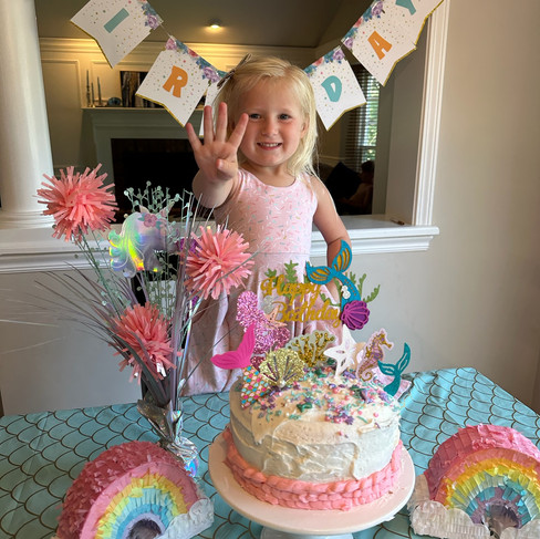 Child holding up four fingers smiles by decorated cake at a birthday party. Festive "Happy Birthday" banner and rainbow decorations visible.