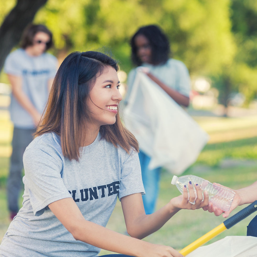 Smiling woman in a "Volunteer" shirt holds a plastic bottle in a sunny park. Two others in the background clean up. Mood: cheerful and collaborative.