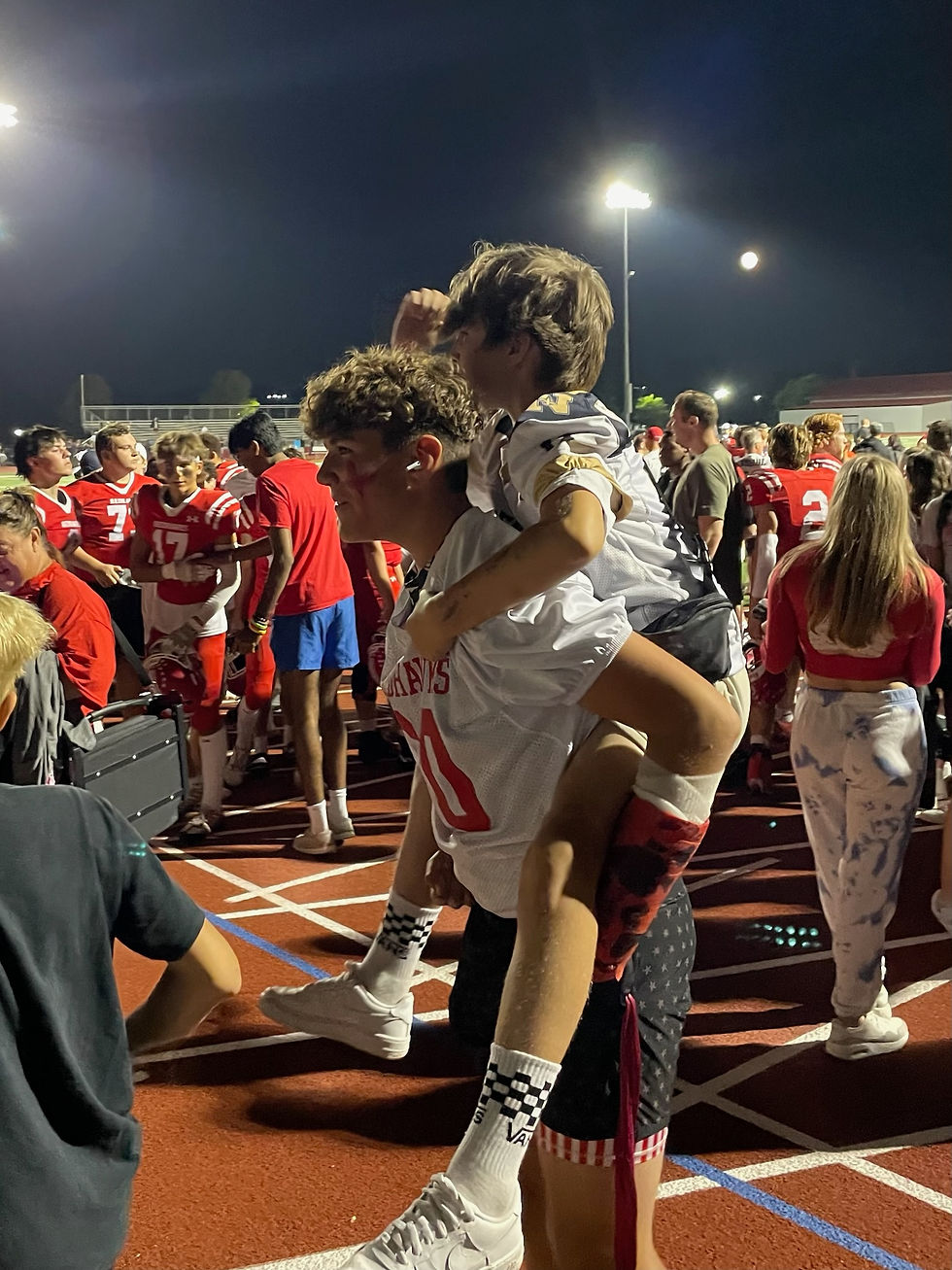 Boy on someone's shoulders at a nighttime sports event. Crowd in red jerseys, vibrant atmosphere. Boy wears checkered socks, looks happy.