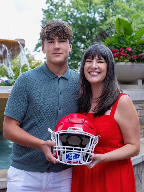 A young man and a woman hold a red football helmet by a fountain. She smiles; both wear red and blue, surrounded by green plants.
