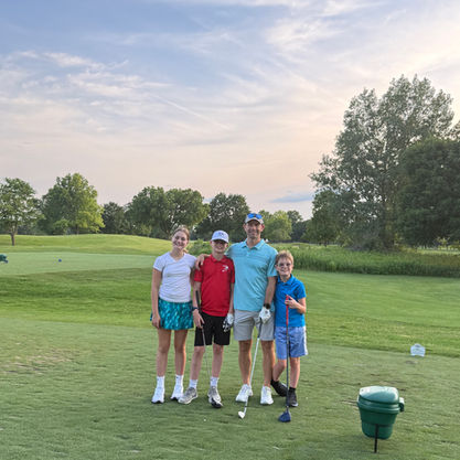 Smiling family of four with golf clubs posing on a green course under a pastel sky.
