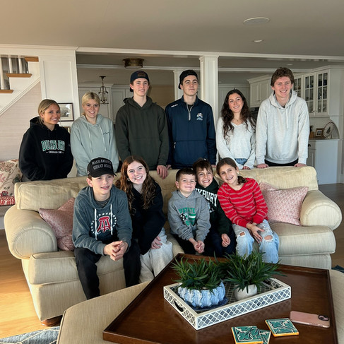 A group of people, mostly seated on beige sofas, smile in a cozy living room. Some wear hoodies. A tray with plants is on the table.